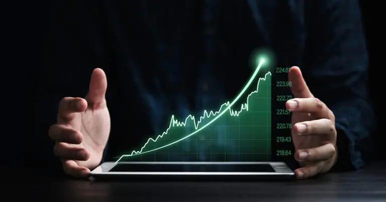 A black shadowy background with two hands emerging out on either side of a digital image of a green, upward trending line chart to signify market growth