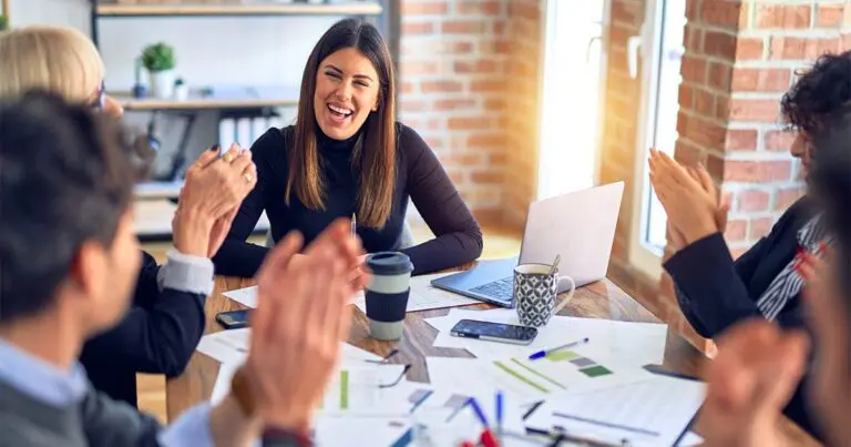 A group of professionals sits around a wooden table in a bright, modern office. A smiling woman in a black top is the center of attention as her colleagues applaud and celebrate her. The table is covered with documents, a laptop, coffee cups, and a smartphone, suggesting a collaborative and productive meeting environment.