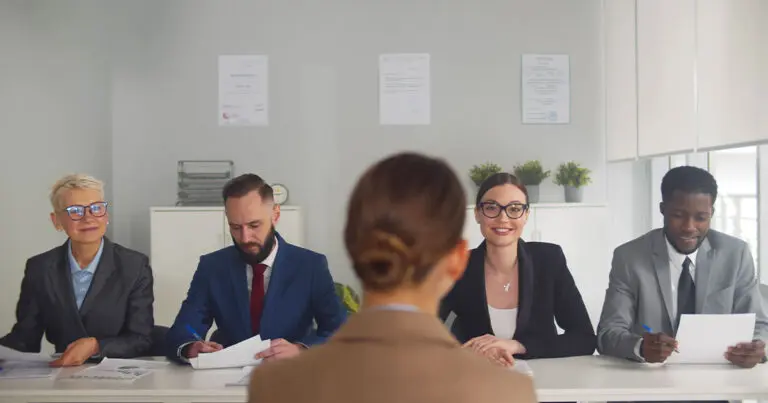 A job candidate sits across from a panel of four interviewers in a brightly lit office. The panel includes two women and two men dressed in business attire, holding papers and pens. One woman is smiling directly at the candidate, while others review documents or prepare to ask questions. The setting is professional, with white walls, potted plants, and certificates on display in the background.