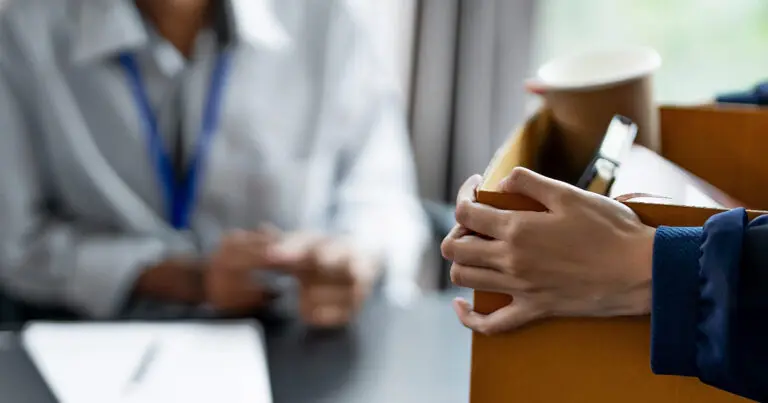 A person holds a cardboard box filled with personal belongings, including a coffee cup and papers, while another person sits across a desk in the background, slightly out of focus. The scene suggests a workplace departure or job transition.