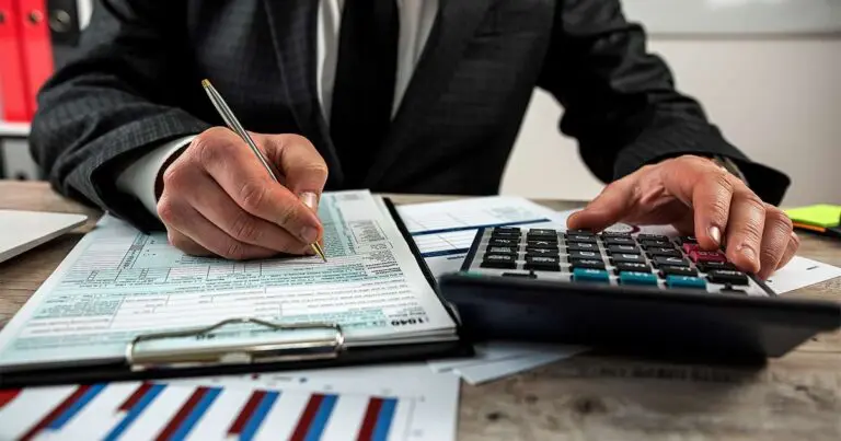 A person in a business suit uses a pen to fill out a financial form on a clipboard while operating a calculator with the other hand. Charts and documents with bar graphs are scattered across the desk, indicating budgeting or financial planning work.