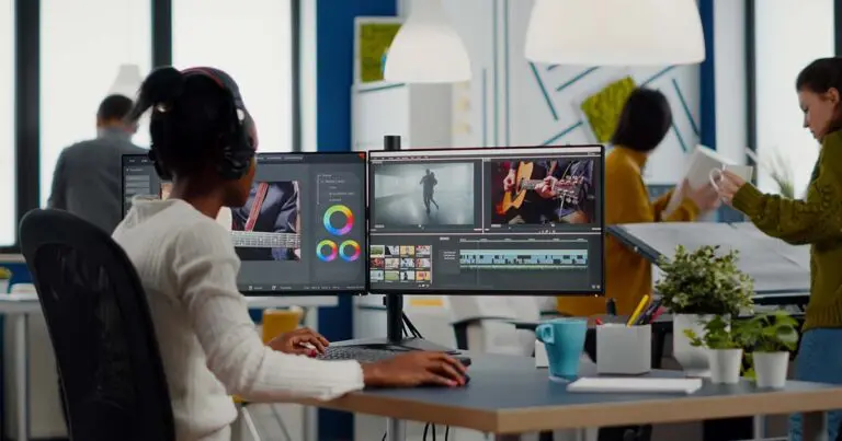 A woman with headphones sits at a dual-monitor setup editing video footage of musicians and dancers in a brightly lit modern office. Color grading tools and timeline editing software are visible. Other professionals work in the background, creating a creative and dynamic environment.