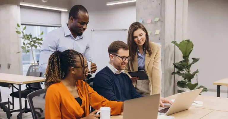 A diverse group of four professionals collaborate around a desk in a modern office. One man sits at a laptop, while a woman beside him gestures toward the screen. Another man stands beside them smiling, and a woman in a blazer holds a tablet, looking on. The setting includes open seating, plants, and a concrete pillar with sticky notes.