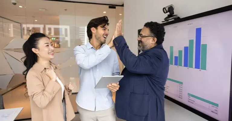 Three professionally dressed colleagues in an office celebrate a success. A man holding a tablet high-fives another man in a blazer while a woman smiles and claps nearby. A large screen in the background displays a bar graph with monthly data.
