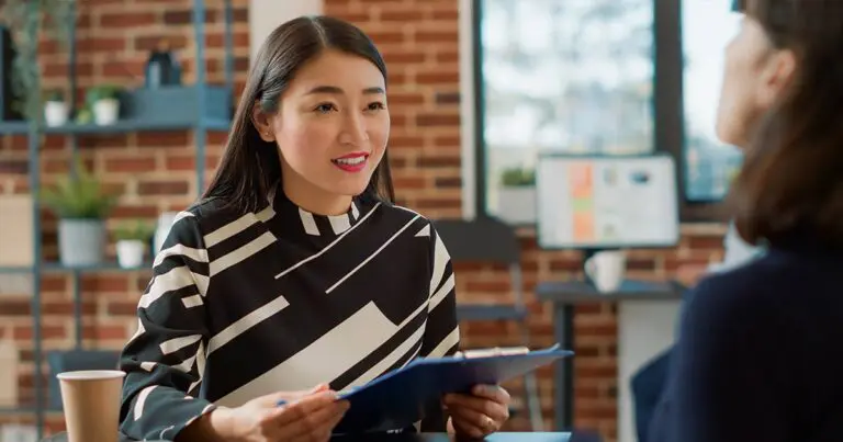 A professional woman with long dark hair, wearing a black and white patterned blouse, holds a clipboard and smiles while interviewing another person in a modern office with exposed brick walls and a computer screen in the background.