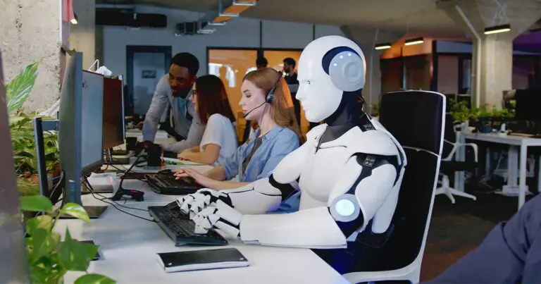 A humanoid robot wearing a headset sits at a desk in a modern office, working at a computer alongside human coworkers. A woman with a headset types on a keyboard next to the robot, while two other colleagues collaborate nearby. The workspace is filled with computers, green plants, and an industrial-style interior with exposed concrete pillars and overhead lighting.