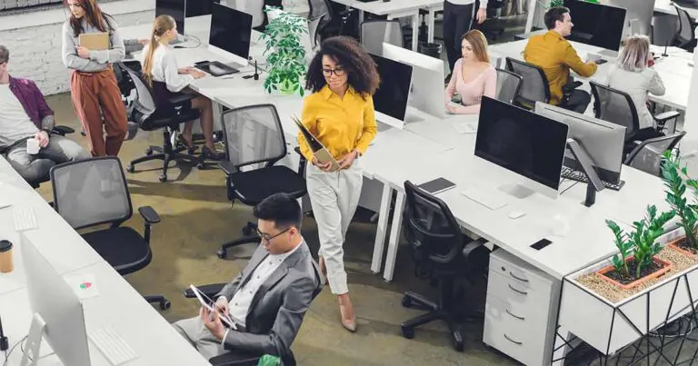 A modern open-plan office with people working at computers and collaborating. A woman in a yellow shirt walks through the workspace holding a tablet, while others sit at desks or talk nearby. The space is bright and organized with white desks, computers, and green plants.