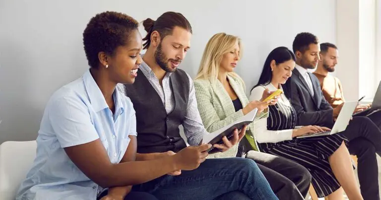 A diverse group of six professionally dressed people sitting in a bright waiting area, each engaged in different activities—reading a document, checking a phone, and working on laptops—suggesting they are waiting for job interviews or a business meeting.