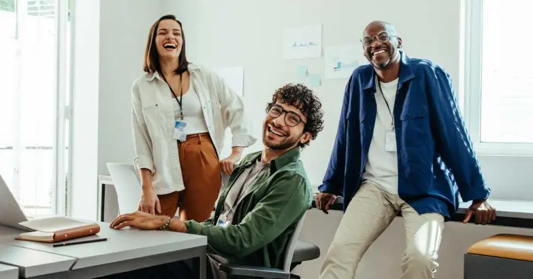 Three coworkers laugh together in a bright, modern office. One person sits at a desk with a laptop while the other two stand nearby, all wearing ID badges and casual clothing. The atmosphere appears friendly and relaxed.