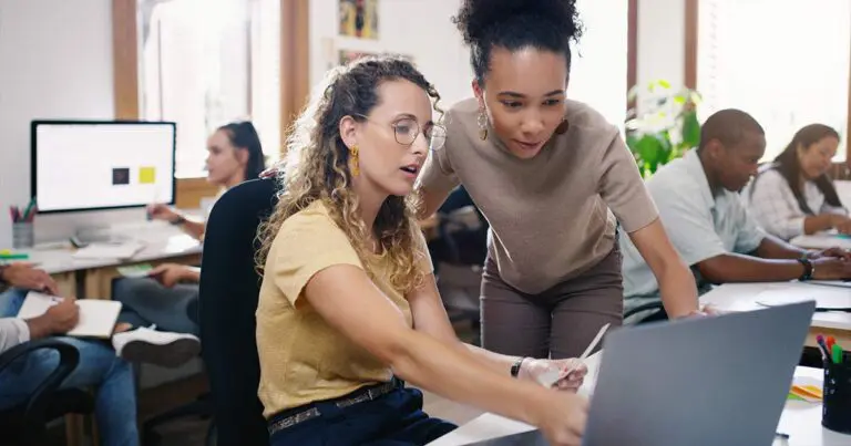 Two colleagues collaborate at a desk, reviewing something on a laptop screen in a bright, modern office. Other team members work in the background, creating a focused and cooperative atmosphere.