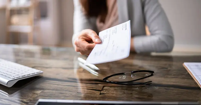 A person in business attire extends a check across a polished desk, with eyeglasses, a keyboard, and paperwork nearby, suggesting a professional or financial transaction.