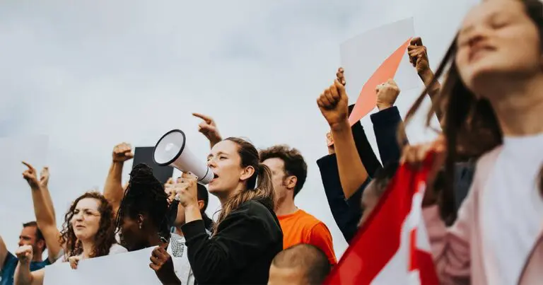 A group of people gathers outdoors holding signs and raising their fists in protest, with one person at the center speaking through a megaphone. The crowd appears energetic and unified under a cloudy sky.