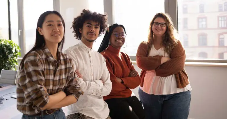 Four people stand together in a bright office space with their arms crossed, looking confidently toward the camera. Sunlight from large windows highlights their relaxed, friendly expressions. The group appears diverse in age, style, and background, suggesting a collaborative or inclusive workplace setting.