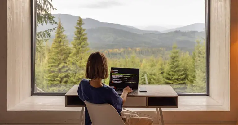 A person sits at a desk facing a large picture window, working on a laptop that displays lines of code. Dense evergreen forest and distant mountain ridges fill the view beyond the glass under an overcast sky. A smartphone rests beside the laptop on the wooden surface.
