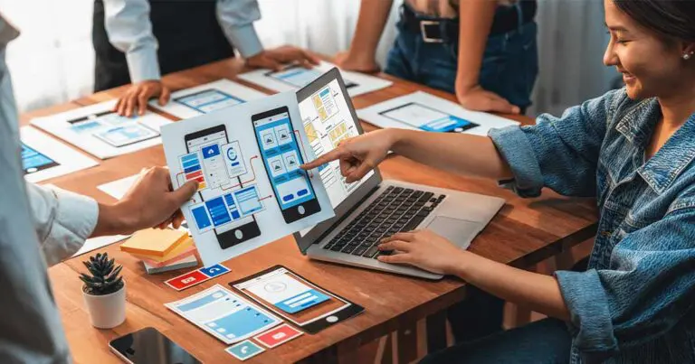 Designers collaborate at a wooden table covered with smartphone interface mockups, pointing to app wireframes displayed on a laptop and printed sheets. The group appears focused on planning or refining a mobile user experience layout.