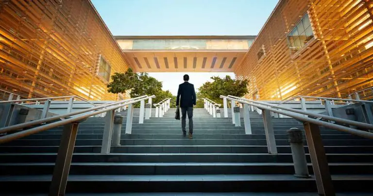 A man in business attire walks up a wide staircase toward a modern building with golden glass walls glowing in the sunlight. The perspective emphasizes the steps and sleek architecture, creating a sense of ambition and forward movement.
