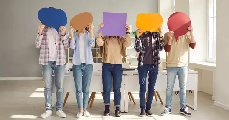 Five people stand in a bright office holding large, colorful speech-bubble cutouts in front of their faces, obscuring their identities. Their casual clothing and the playful props create a light, collaborative atmosphere.