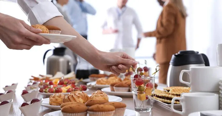 A hand reaches across a buffet table to pick up a skewer with grapes, cheese, and olives beside trays of pastries, muffins, and waffles. Coffee carafes and stacked cups sit nearby, while a small group of people talk in the blurred background.
