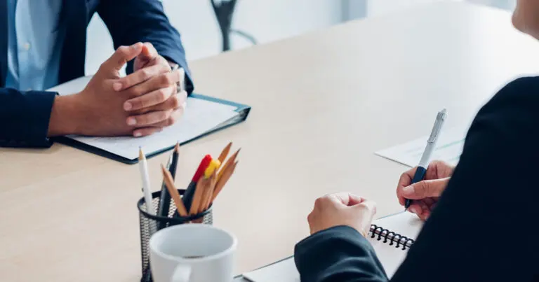 Two people sit across a table in a professional setting, with one person resting clasped hands on a folder while the other holds a pen over an open notebook. A cup, papers, and a container of pencils are on the table, suggesting a formal conversation or interview.