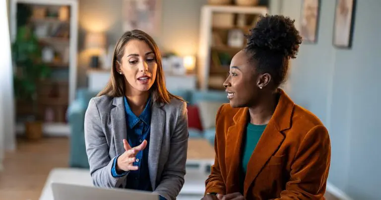 Two women sit side by side in a comfortable office setting, discussing something on a laptop as one gestures while speaking. Their engaged expressions and professional clothing suggest a focused, collaborative conversation.