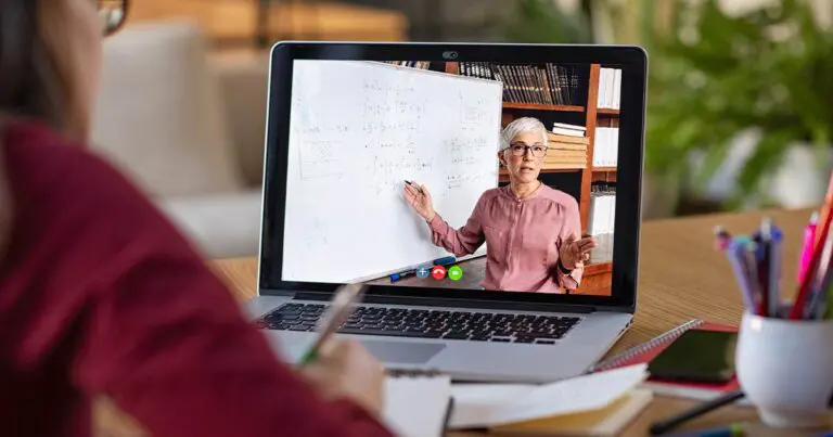 A student takes notes while watching a virtual lesson on a laptop showing an instructor standing beside a whiteboard covered in mathematical formulas and graphs. The instructor gestures toward the equations as she explains them during the video call.