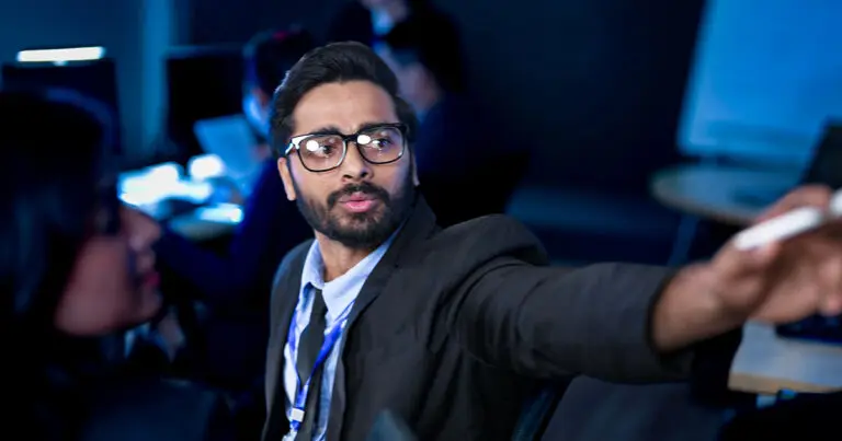 A professional wearing glasses and a suit gestures toward a screen while speaking in a dimly lit office, with coworkers working at computers in the background. The scene conveys active collaboration and decision-making in a data or product-focused team environment.