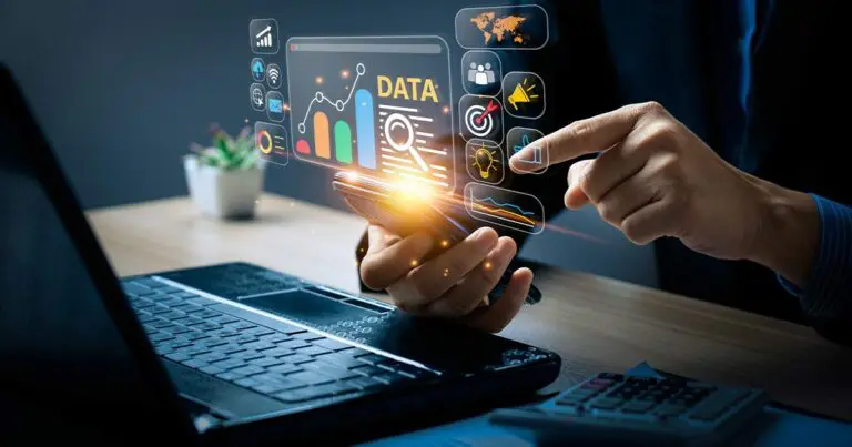 A person holds a smartphone above a desk while tapping floating digital dashboard icons labeled “DATA,” with charts, graphs, and analytics symbols glowing in midair. A laptop keyboard and calculator sit nearby, reinforcing a work setting focused on data analysis and performance tracking.