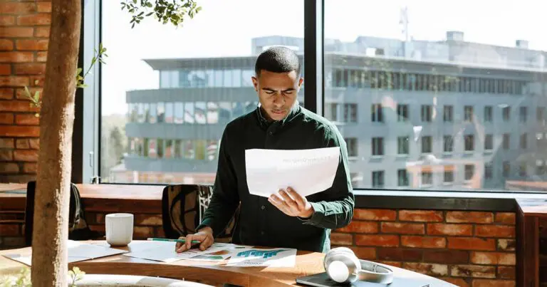 A man stands at a wooden desk in a sunlit office, studying printed documents with charts and text while holding a pen. Large windows behind him reveal modern office buildings, and a coffee mug and headphones rest on the desk, suggesting quiet talent doing focused, analytical work.