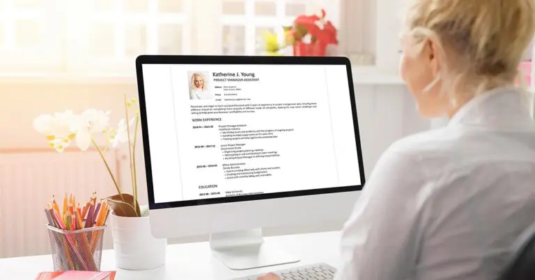 A person sits at a desk looking at a computer monitor displaying a résumé titled “Katherine J. Young, Project Manager Assistant.” The screen shows a professional headshot and sections for work experience and education in a clean, minimalist layout, with office supplies and a plant visible in the workspace.