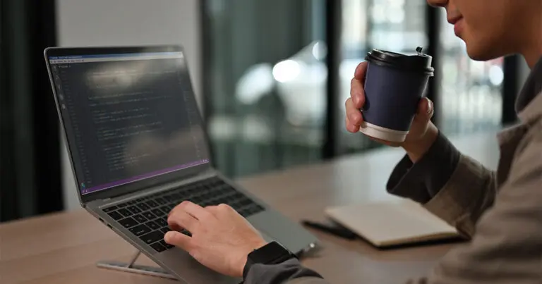 Person working at a laptop displaying lines of code, holding a takeaway coffee cup at a desk with a notebook in a modern office setting. The scene conveys focused, informal software development work, aligning with the theme of AI-assisted or vibe coding.