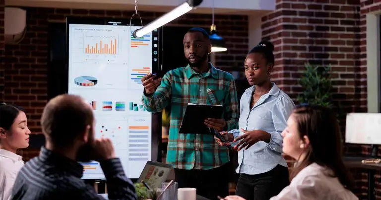 A diverse group of professionals stands in a modern office, with two people presenting data from a large digital screen showing charts, graphs, and dashboards. The scene conveys a focused discussion around data-driven decision-making, collaboration, and the human expertise behind advanced technology work