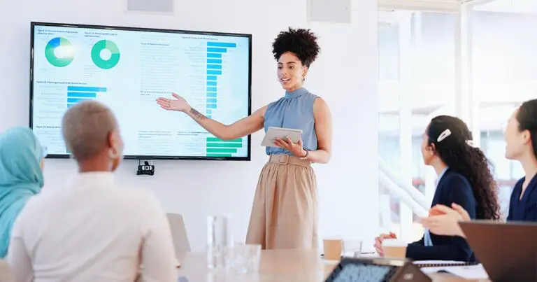 A presenter stands at the front of a conference room, gesturing toward a large screen displaying charts and data while holding a tablet. Several attendees sit around a table listening, suggesting a professional meeting or data-driven storytelling.