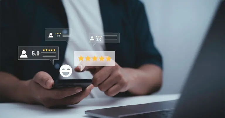 Hands hold a smartphone above a desk while digital review icons float on screen, including five-star ratings and numeric scores like 5.0, 4.0, and 3.0. A smiling face icon and star graphics suggest customer feedback or satisfaction, with a laptop blurred in the background to imply an online or workplace setting.