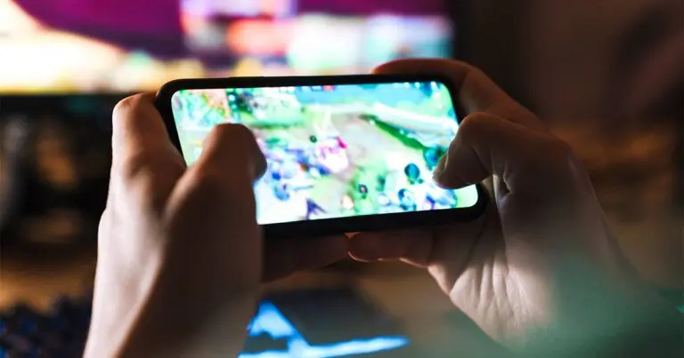 Hands hold a smartphone in landscape orientation while a colorful, fast-moving game plays on the screen, with thumbs actively controlling the action. The background is dim and out of focus, emphasizing mobile gaming and in-game monetization in a low-light setting.