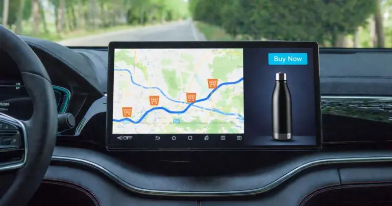 A car’s dashboard display shows a navigation map with a driving route marked by multiple shopping cart icons along the way. To the right of the map, a digital ad panel displays a black water bottle with the visible text “Buy Now,” suggesting in-car advertising integrated with navigation.