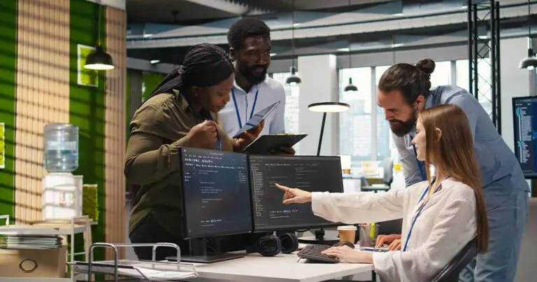 Five junior developers collaborate around a desk in a modern office, reviewing code on dual monitors while one person points at the screen and others take notes on tablets. The scene conveys teamwork, mentorship, and hands-on problem-solving in a tech workplace, reflecting the continued importance of entry-level developers alongside AI tools.
