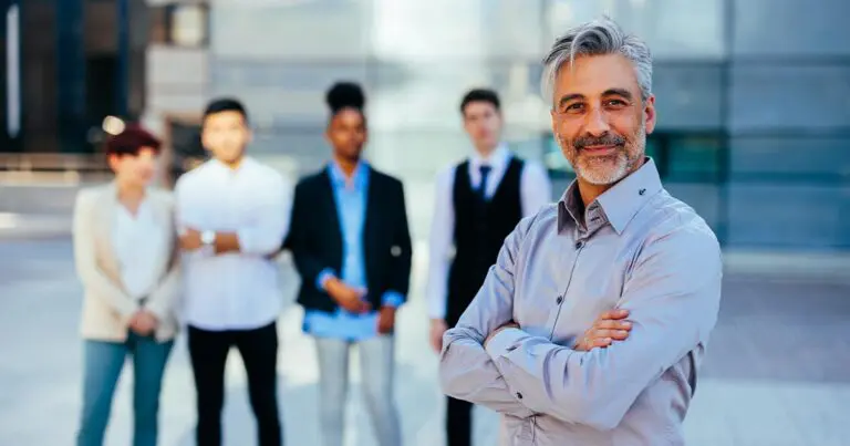 A middle-aged professional with gray hair stands confidently with arms crossed in the foreground, smiling at the camera, while a diverse group of colleagues stands slightly out of focus behind him outside a modern office building. The composition emphasizes leadership and generational transition in a contemporary workplace setting.