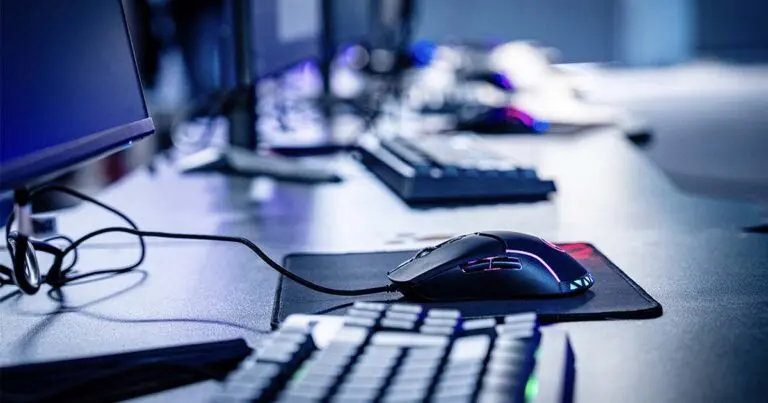 Close-up of a desktop gaming setup with a wired mouse glowing in neon colors on a mousepad, surrounded by a keyboard and monitors. The scene is softly lit with cool blue and purple tones, creating a focused, tech-oriented atmosphere.