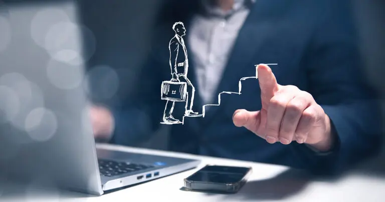 A person in a business suit points toward a laptop screen displaying a white line drawing of a man with a briefcase climbing upward steps. The scene suggests career advancement or professional growth, with a smartphone resting on the desk beside the laptop.