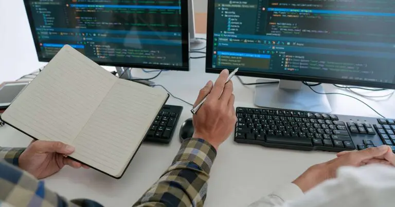 Two off camera people sit at a computer desk in front of two monitors and keyboards. The person on the left holds a notebook and points toward one of the monitors that has design code on the screen to symbolize the gap between designers and developers