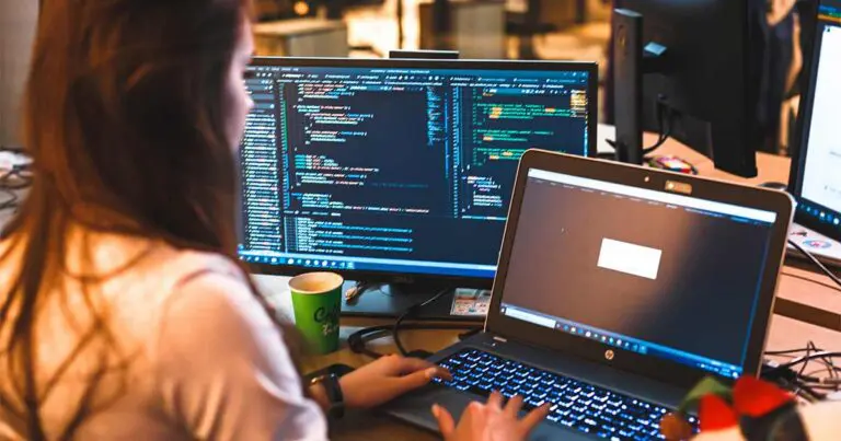 A woman sits at a desk with her back to the camera facing two computer monitors both containing code and code notifications to represent ai productivity and ai coding tools