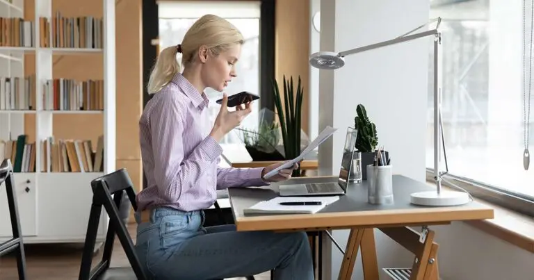 A blonde-haired woman sits in a naturally lit room at a wooden desk in front of a large picture window. She is wearing blue jeans and a pink button up and is speaking into her phone while she checks notes on a legal pad to represent how conversational AI is affecting customer experience