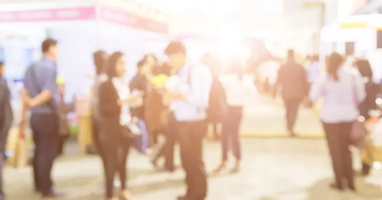A blurry photo of a brightly lit room where various individuals dressed in business casual attire stand around ot represent a job fair or the job market.
