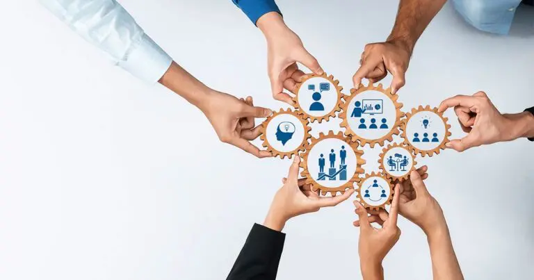 Six arms reach in from out of frame holding and interconnecting wooden cogs with symbols like the side profile of a head, profiles of people standing on steps, etc to represent scenario based workforce planning