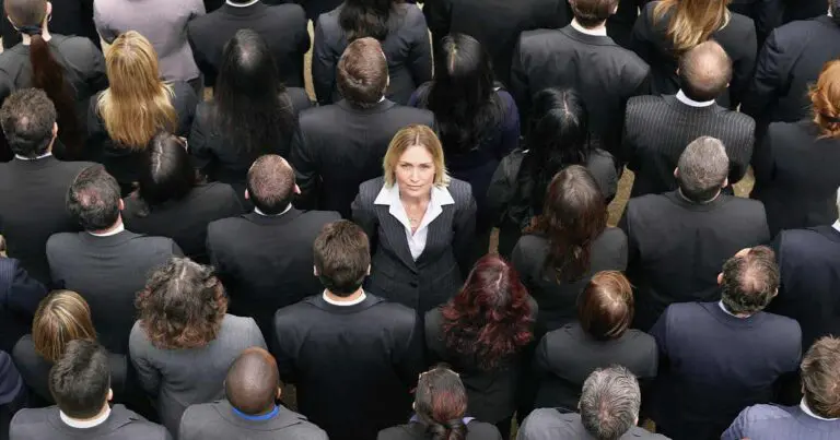 A top-down photo of a group of people wearing black or gray suits all facing away from the camera except for one woman in the center facing the opposite direction and looking up at the camera