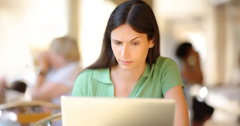 A dark haired woman in a green collard t-shirt sits in front of a laptop and looks quizzically at the screen to represent ghost job postings