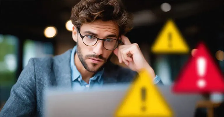 Concerned man wearing glasses looks at a laptop screen while resting his head on his hand, suggesting worry or concentration. Blurred warning icons with exclamation marks in the foreground reinforce a theme of security risks and a SaaS supply chain breach.