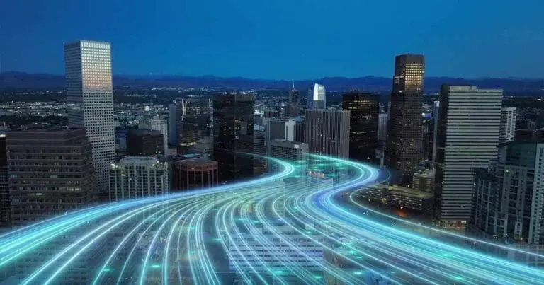 Aerial view of a downtown city skyline at dusk, with illuminated buildings against a deep blue sky. Bright, glowing light trails curve through the streets below, suggesting fast-moving traffic or digital data flow. The scene conveys a futuristic, high-tech urban environment.