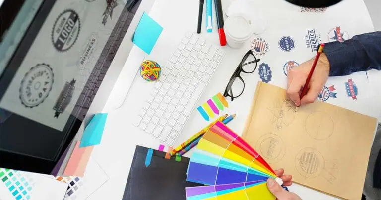 Overhead view of a designer’s workspace with a computer, keyboard, and colorful swatches spread across the desk while a hand sketches logo concepts in a notebook. Sticky notes, pens, and printed badge-style graphics labeled “Made in America” and “Vote” suggest branding and handmade design work.