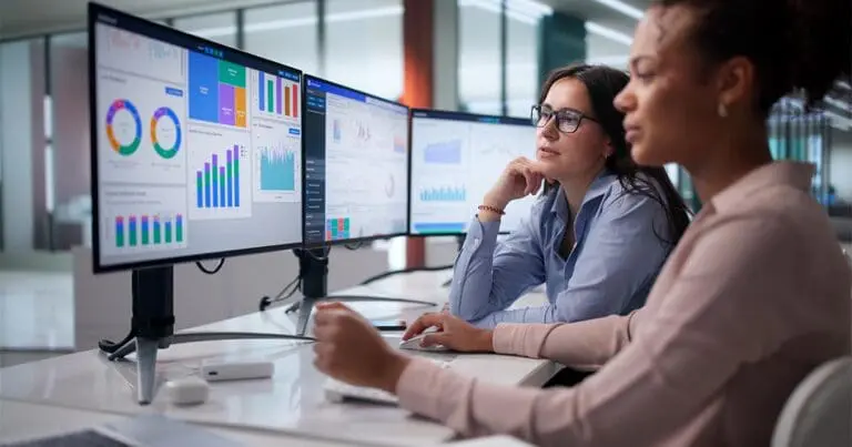 Two professionals sit side by side at a desk, studying multiple large monitors displaying colorful data dashboards with charts, graphs, and performance metrics. Their focused expressions and collaborative posture suggest analysis of marketing data, aligning with AI-driven performance strategy.